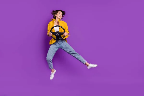 Foto de impressionado engraçado jovem mulher vestida camisa amarela sorrindo pulando alto segurando volante isolado cor roxa fundo — Fotografia de Stock