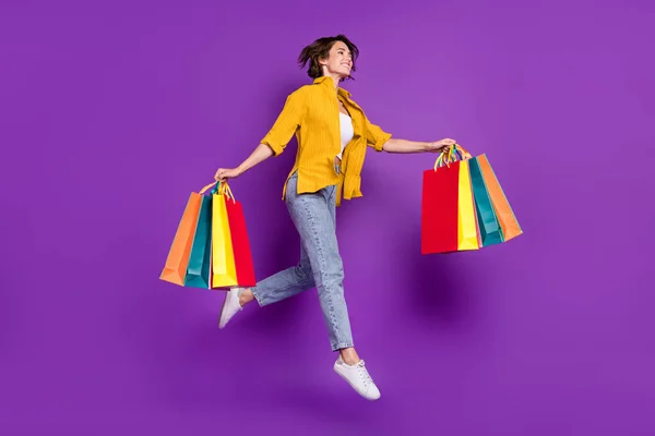 Foto van funky mooie jonge dame dragen geel shirt springen hoge holding shopping tassen op zoek naar lege ruimte glimlachen geïsoleerde violette kleur achtergrond — Stockfoto