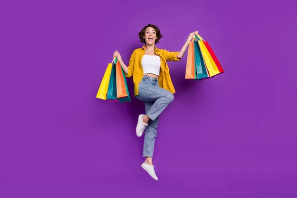 Photo of funny impressed young woman dressed yellow shirt smiling jumping high holding bargains isolated purple color background