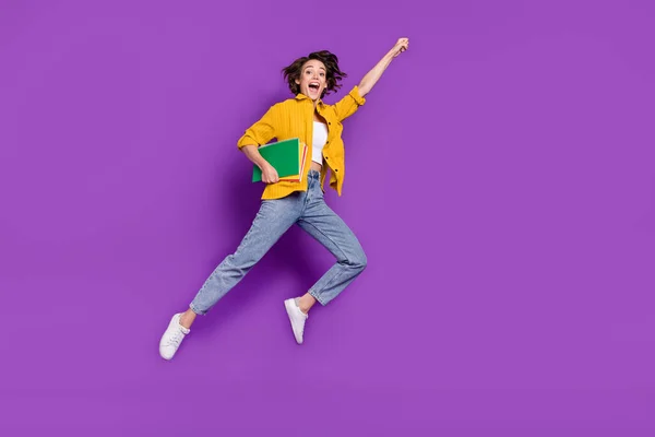 Foto de animado engraçado jovem mulher vestida camisa amarela saltando alta segurando copybooks levantando punho isolado cor roxa fundo — Fotografia de Stock