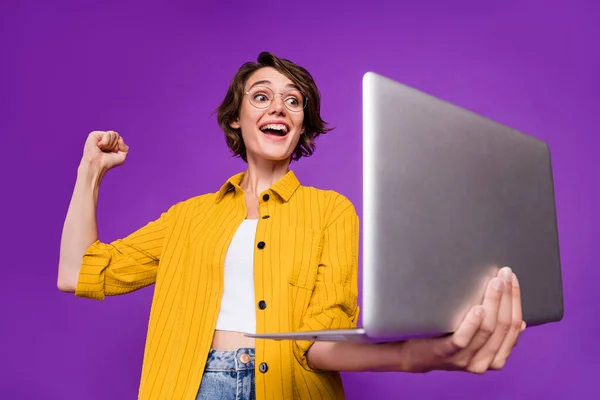 Photo of sweet lucky young woman dressed yellow shirt glasses looking modern device rising fist isolated purple color background — Stock Photo, Image