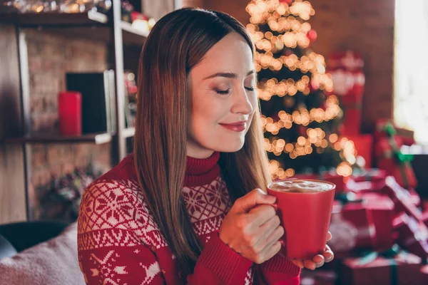 Фото мрійливої чарівної зрілої леді в червоному пуловері, що посміхається насолоджуючись різдвяним напоєм в приміщенні домашньої кімнати будинку — стокове фото