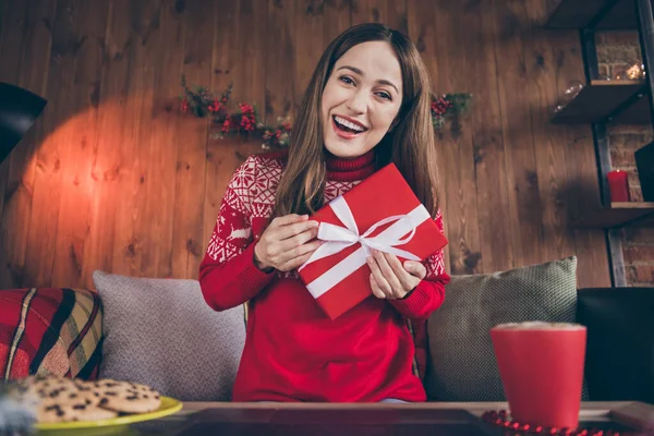 Foto av glad drömmande ung kvinna klädd röd tröja kommunicerar modern gadget håller nytt år gåva inomhus rum hem hus — Stockfoto