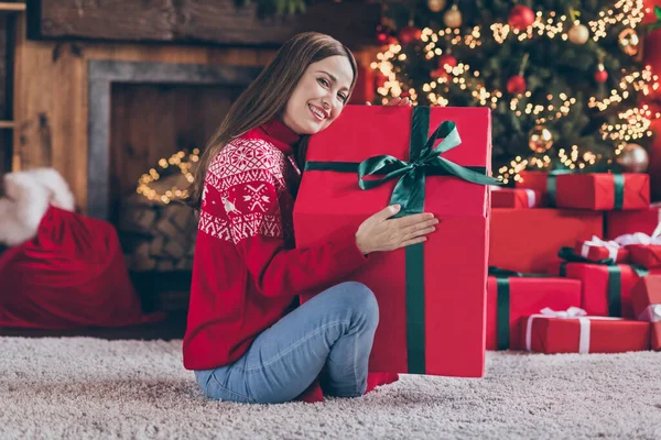 Фото довольно мечтательная молодая женщина одета красный свитер обнимая новый год большой подарочный коробок улыбаясь в помещении комната домашний дом — стоковое фото