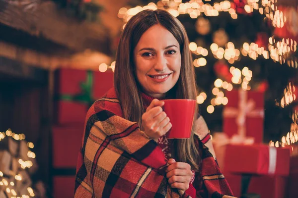Φωτογραφία από θετική γλυκιά ώριμη κυρία φορούν κόκκινο πουλόβερ χαμογελώντας απολαμβάνοντας χριστουγεννιάτικο ποτό σε εσωτερικούς χώρους σπίτι δωμάτιο — Φωτογραφία Αρχείου