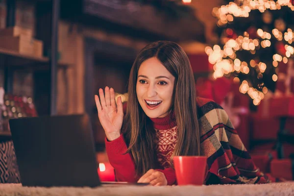 Foto av glad söt ung kvinna klädd röd tröja talar modern gadget skicka nytt år hälsning viftande arm inomhus rum hem hus — Stockfoto