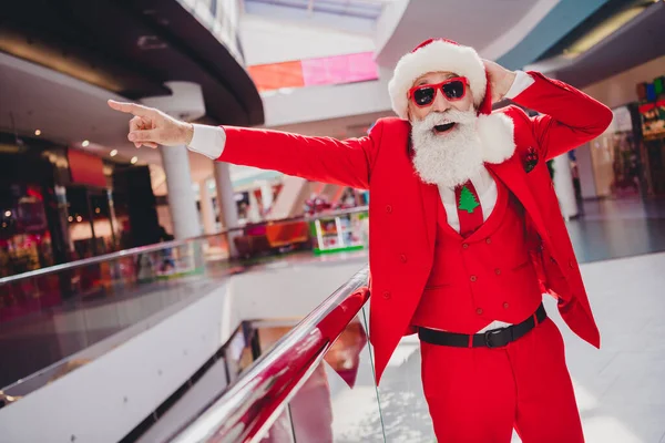 Photo of fashion trendy impresja Santa Claus nosić ciemne okulary wskazując palec daleko uśmiechnięty kryty sklep butikowy — Zdjęcie stockowe