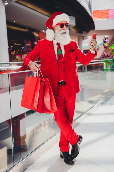 Vertical comprimento total corpo tamanho vista de atraente barbudo alegre Papai Noel comprar presentes usando aplicativo ordem gadget no shopping center dentro de casa — Fotografia de Stock