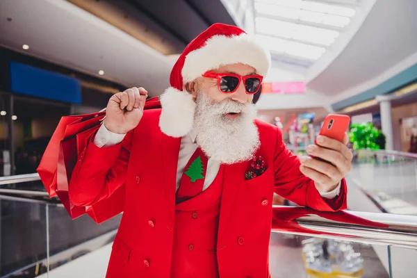 Foto de Santa hold telefone transportar sacos presentes fazer social mídia post desgaste chapéu de vidro vermelho smoking no centro comercial supermercado — Fotografia de Stock