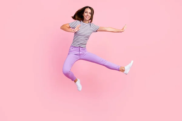 Фото чарівної сильної молодої леді в смугастому вбранні, посміхаючись стрибає з високою практикою карате ізольований рожевий кольоровий фон — стокове фото