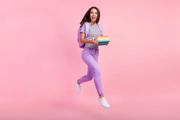 Foto de brillante emocionado joven dama usar traje a rayas mochila sonriendo celebración copybooks saltar alto aislado color rosa fondo — Foto de Stock
