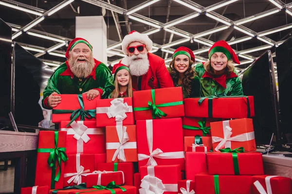 Çekici noel baba asistanlarının fotoğrafı. Gülümseyen kostümler giyerler. Bir sürü hediye kutusunu kapalı bir alışveriş merkezinde tutarlar. — Stok fotoğraf