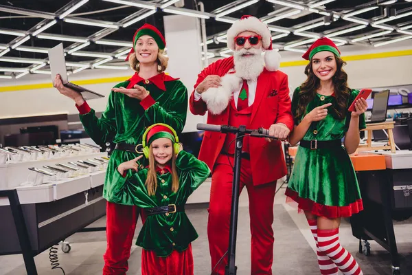 照片上滑稽漂亮的圣诞老人助手们穿着服装微笑向您推荐室内现代电子产品购物中心 — 图库照片