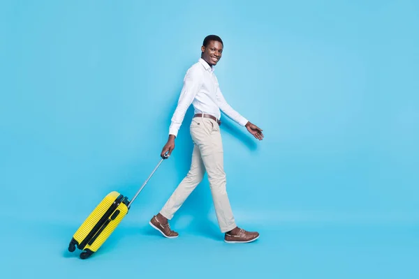 Φωτογραφία του θετικού αρκετά σκούρο δέρμα τύπος ντυμένος επίσημη πουκάμισο περπάτημα κρατώντας αποσκευές χαμογελώντας απομονωμένο φόντο μπλε χρώμα — Φωτογραφία Αρχείου