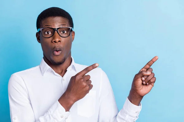 Foto de impressionado chocado pele escura homem desgaste branco camisa óculos apontando vazio espaço isolado azul cor fundo — Fotografia de Stock