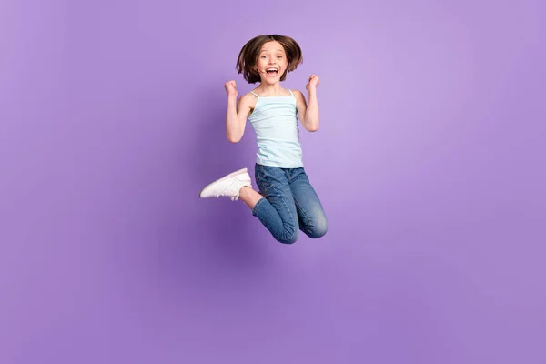 Ganzkörperfoto von niedlichen braunen Frisur kleine Mädchen springen Hände Fäuste tragen blaue Top Jeans isoliert auf violettem Hintergrund — Stockfoto