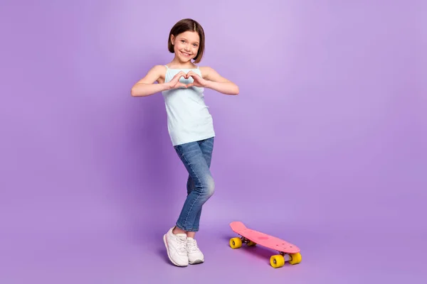 Foto de comprimento total de penteado marrom doce menina pequena mostrar coração desgaste azul top jeans perto skate isolado no fundo cor violeta — Fotografia de Stock