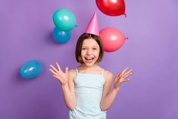 Foto di allegro felice positivo piccola ragazza caduta aria palloncini sorriso indossare cappello cono isolato su sfondo di colore viola — Foto Stock