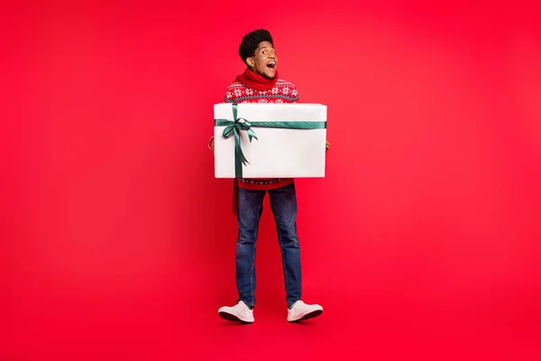 Ganzkörper-Foto von Millennial Brunet Kerl halten Geschenk tragen Urlaub Schal Pullover Jeans Schuhe isoliert auf rotem Hintergrund — Stockfoto