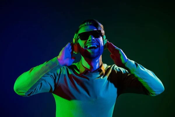 Φωτογραφία του νεαρού δροσερό τύπος ακούσετε μουσική φορούν ακουστικά γυαλιά πουλόβερ απομονώνονται σε σκούρο μπλε φόντο χρώμα — Φωτογραφία Αρχείου