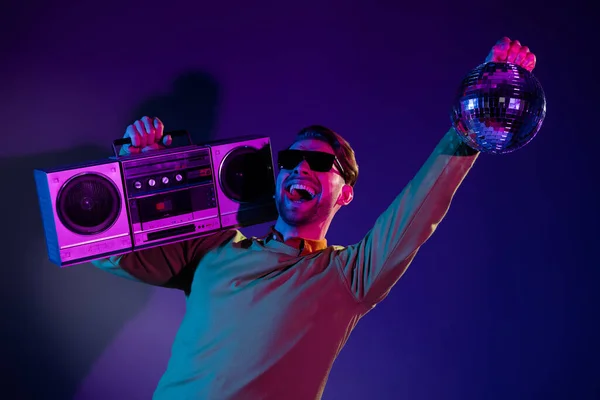 Fotografie veselý mladý cool šťastný muž držet ruku boombox disco míč clubber izolované na neonovém pozadí — Stock fotografie