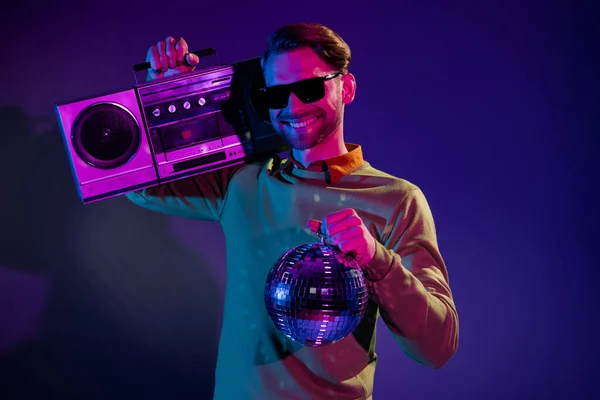 Foto retrato joven en gafas de sol sonriendo manteniendo bola disco grabadora de cinta retro gradiente aislado color violeta fondo — Foto de Stock