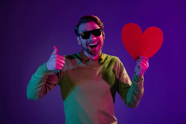 Foto de jovem feliz positivo segurar mão coração de papel vermelho fazer thumb-up isolado no fundo de cor de néon — Fotografia de Stock