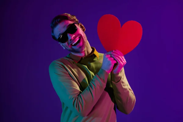 Retrato fotográfico del hombre con gafas de sol mostrando una postal roja en forma de corazón aislada sobre fondo de color púrpura — Foto de Stock