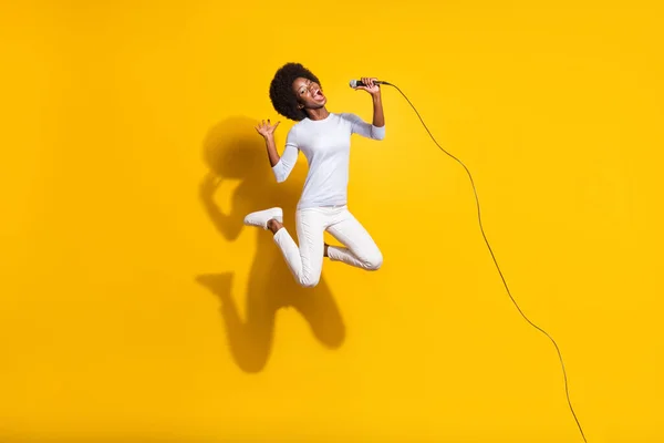 フル長さ写真の暗い肌の女性ジャンプアップ歌うミックウェアホワイトパンツプルオーバー隔離された黄色の色の背景 — ストック写真
