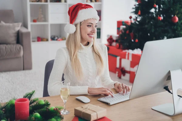 Foto av ung positiv lycklig kvinna bär tomte klo hatt sitta skrivbord skriva pc jul inomhus hus — Stockfoto