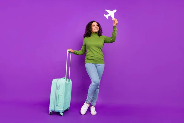 Foto de senhora africana sonhadora brilhante vestida de gola alta verde sorrindo segurando bagagem de avião branco fundo cor violeta isolado — Fotografia de Stock