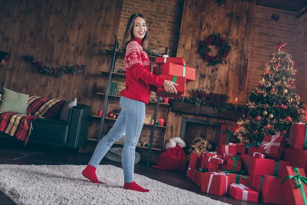 Kaygısız, çekici bir genç bayanın fotoğrafı kırmızı bir kazak giyip gülümseyerek elinde Noel hediyeleriyle evin içinde. — Stok fotoğraf