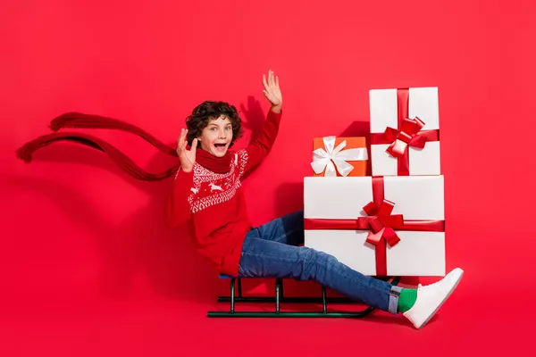 Foto von unbeschwert froh Junge sitzen Schlitten liefern Stapel Geschenkbox tragen Hirsch Schmuck Pullover Schal isoliert rote Farbe Hintergrund — Stockfoto