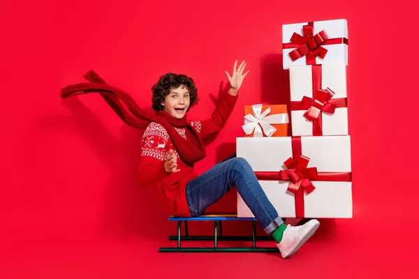 Foto av ryttare rolig pojke sitta släde bära stack presentbox bära rådjur prydnad pullover halsduk isolerad röd färg bakgrund — Stockfoto
