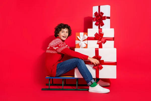 Foto von erstaunt schockiert Junge sitzen Schlitten tragen Haufen Box tragen Hirsch Ornament Pullover isoliert rote Farbe Hintergrund — Stockfoto
