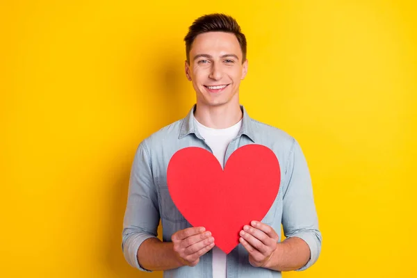 Retrato de chico alegre atractivo sosteniendo en las manos tarjeta de corazón grande aislado sobre fondo de color amarillo brillante — Foto de Stock