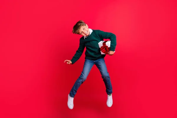 Foto av roliga söta skolpojke klädd grön tröja hoppa högt innehav presentförpackning ler isolerad röd färg bakgrund — Stockfoto