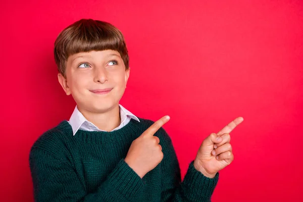 Foto de niño de escuela lindo de ensueño usar jersey de punto sonriente apuntando buscando espacio vacío aislado color rojo fondo —  Fotos de Stock