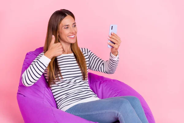 Foto av glänsande drömmande dam bära randig tröja sittande bönpåse visar tummen upp tackning selfie modern gadget isolerad rosa färg bakgrund — Stockfoto