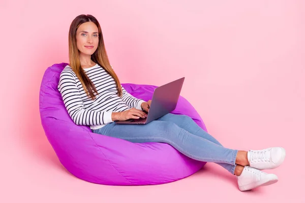 美しいです光沢のある若い女性の服を着た縞模様プルオーバー座っている豆の袋作業現代的なデバイス笑顔隔離されたピンク色の背景 — ストック写真