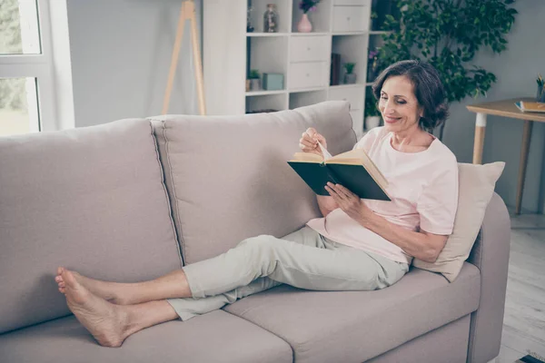 Фото мечтательной милой пожилой леди носить случайный наряд улыбаясь сидя диван чтение книги в помещении комнате дом — стоковое фото