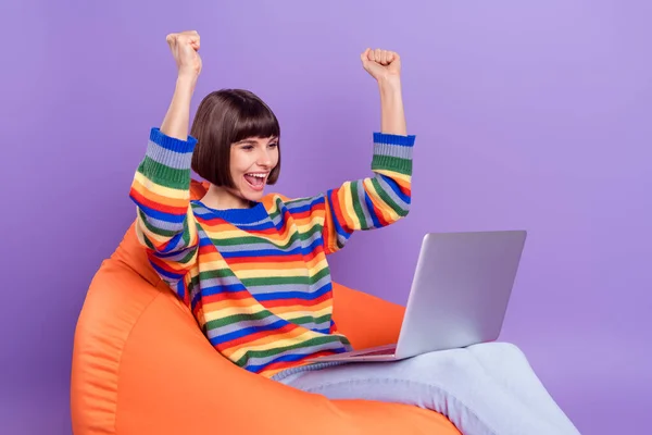 Foto de la señora asombrada sentarse beanbag celebrar netbook levantar los puños usar jersey a rayas aislado color violeta fondo —  Fotos de Stock