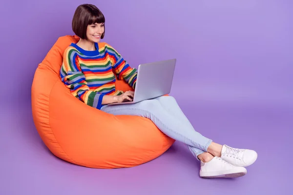 Foto wanita ceria duduk beanbag memegang netbook tampilan layar memakai bergaris pullover terisolasi warna ungu latar belakang — Stok Foto