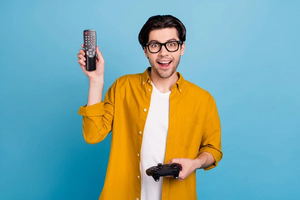 Fotografie vzrušený funky nerd chlap držet dálkové ovládání joystick nosit brýle žlutá košile izolované modré barvy pozadí — Stock fotografie