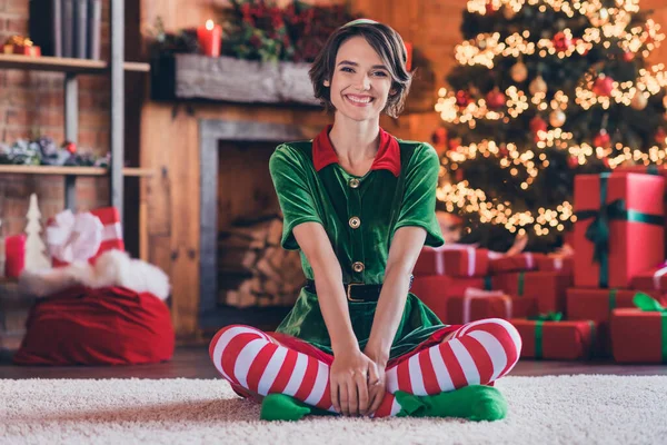 Фото милої смішної молодої жінки, одягненої в зелений костюм, посміхаючись святкувати новий рік, сидячі ноги схрещені в приміщенні будинку — стокове фото
