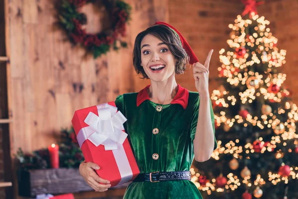 Фото солодкої розумної молодої леді в одязі ельфа, що тримає подарунок святкування Різдва вказуючи палець посміхаючись в приміщенні домашньої кімнати будинку — стокове фото