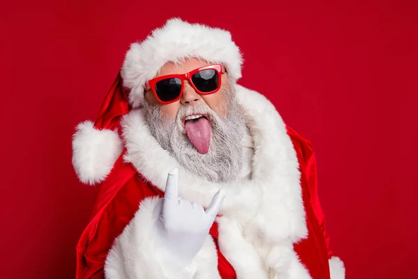 Bild av cool funky Santa claus bära röd kostym hatt mörka glasögon visar hårt rock tecken tunga isolerad röd färg bakgrund — Stockfoto