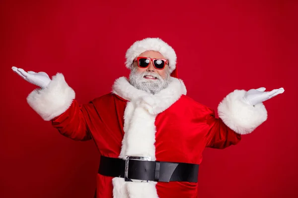 Zdjęcie wesoły człowiek dwie ręce demonstrują puste warianty przestrzeni nosić Santa kapelusz kostium izolowany czerwony kolor tła — Zdjęcie stockowe