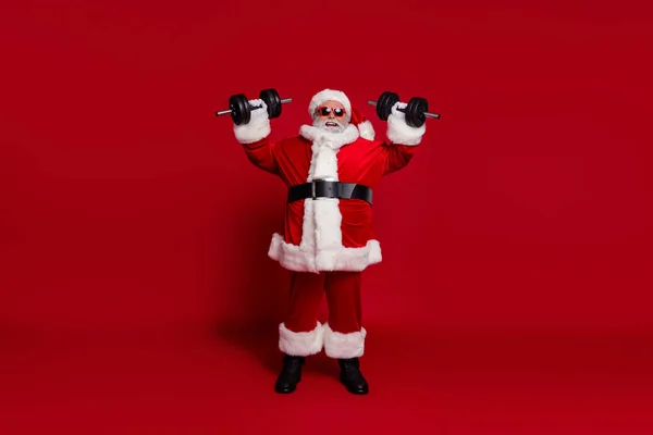 Foto de sporty ativo enérgico homem velho fazer exercício de levantamento de peso desgaste santa chapéu traje isolado cor vermelha fundo — Fotografia de Stock