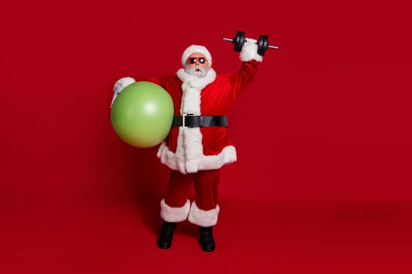 Foto av galen gammal man hålla passform boll lyft tunga x-mas vikter bära Santa hat kostym isolerad röd färg bakgrund — Stockfoto
